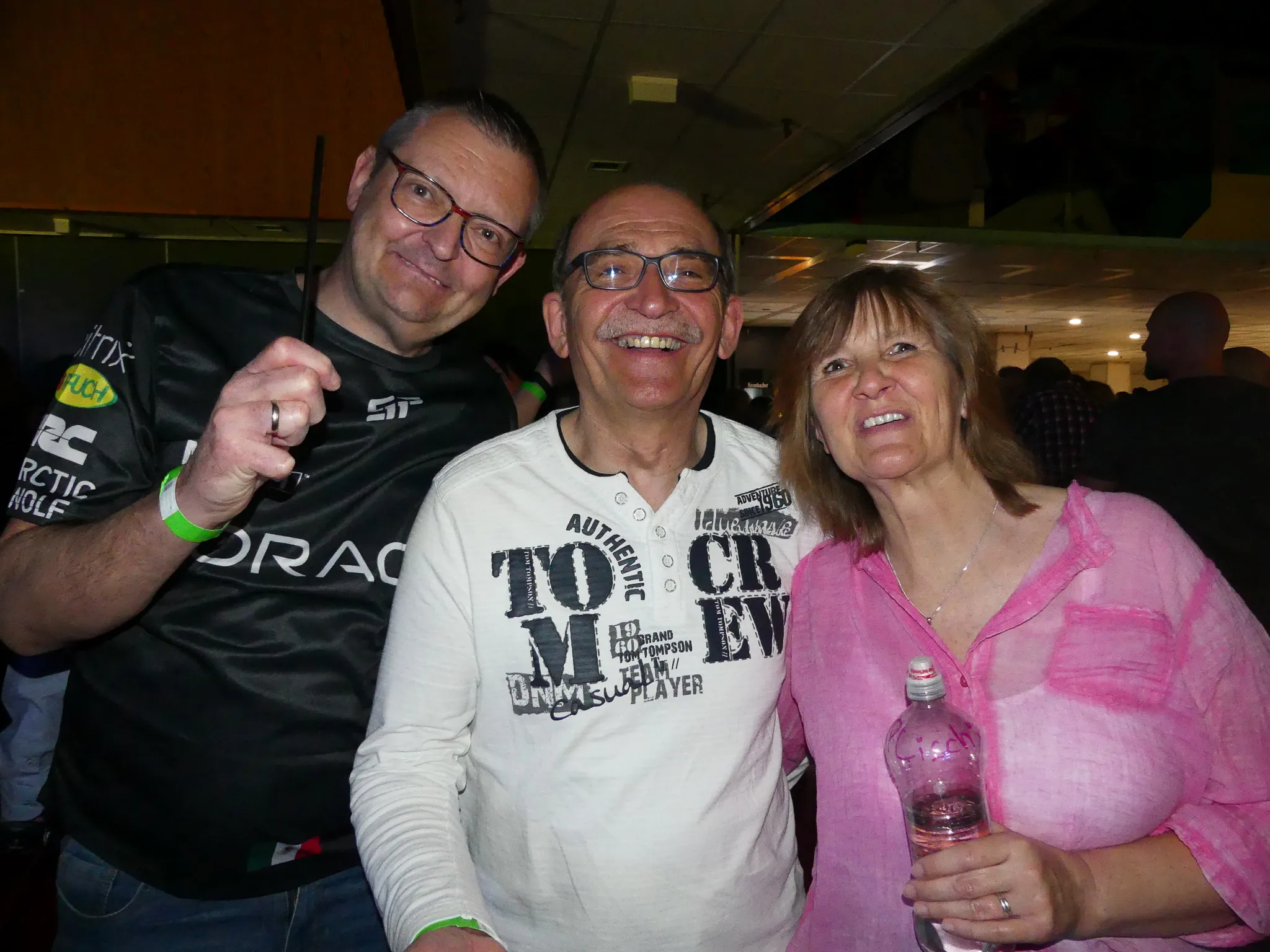 Drei Personen lächeln in die Kamera. Links ein Mann mit Brille und schwarzem Shirt, in der Mitte ein Mann mit gemustertem Shirt und rechts eine Frau mit rosa Oberteil. Im Hintergrund sind weitere tanzende Menschen zu sehen.