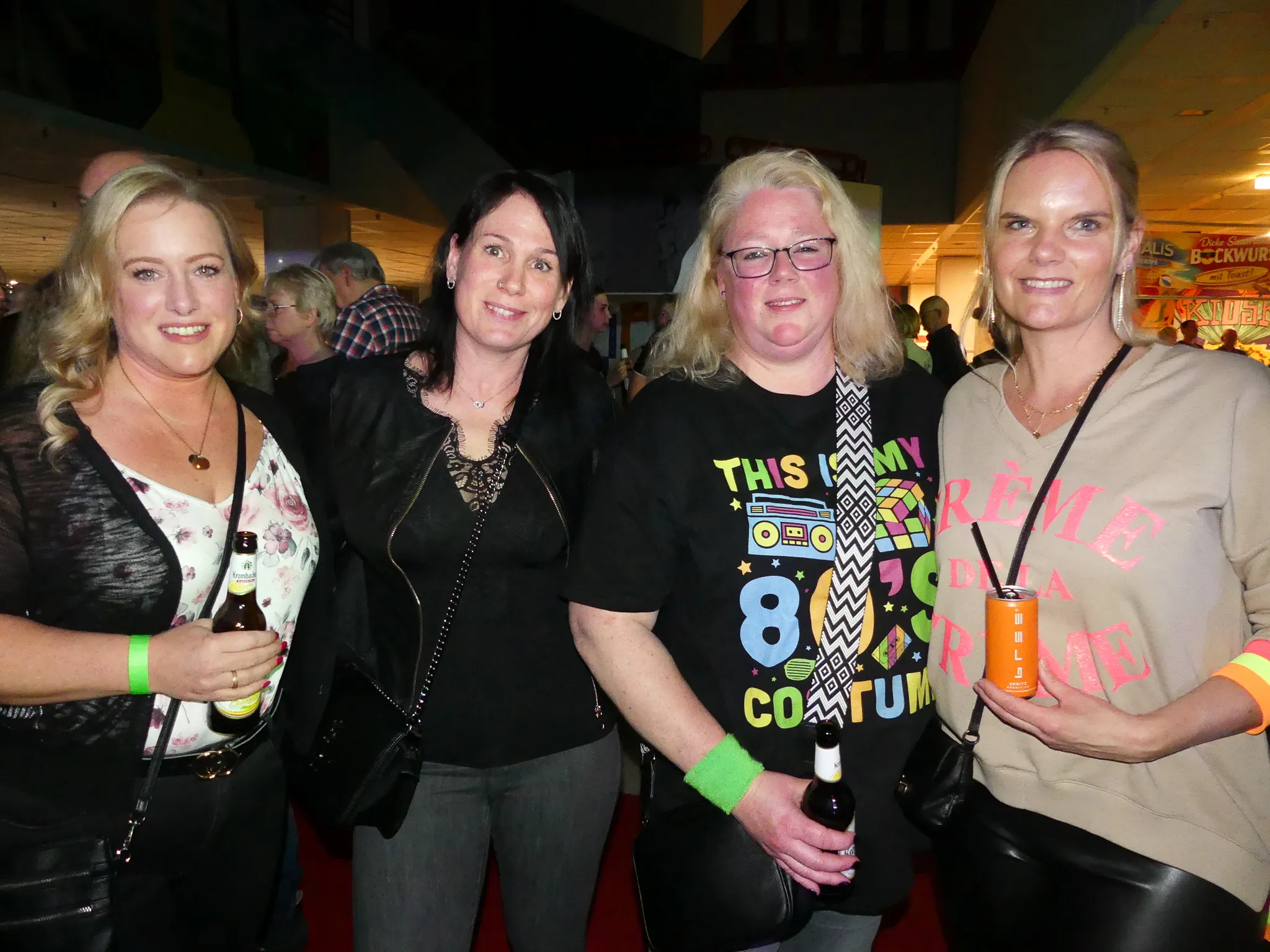 Vier lächelnde Frauen stehen eng zusammen, zwei von ihnen halten Getränke. Die Frau in der Mitte trägt ein T-Shirt mit dem Aufdruck „This is my 80s Costume“.