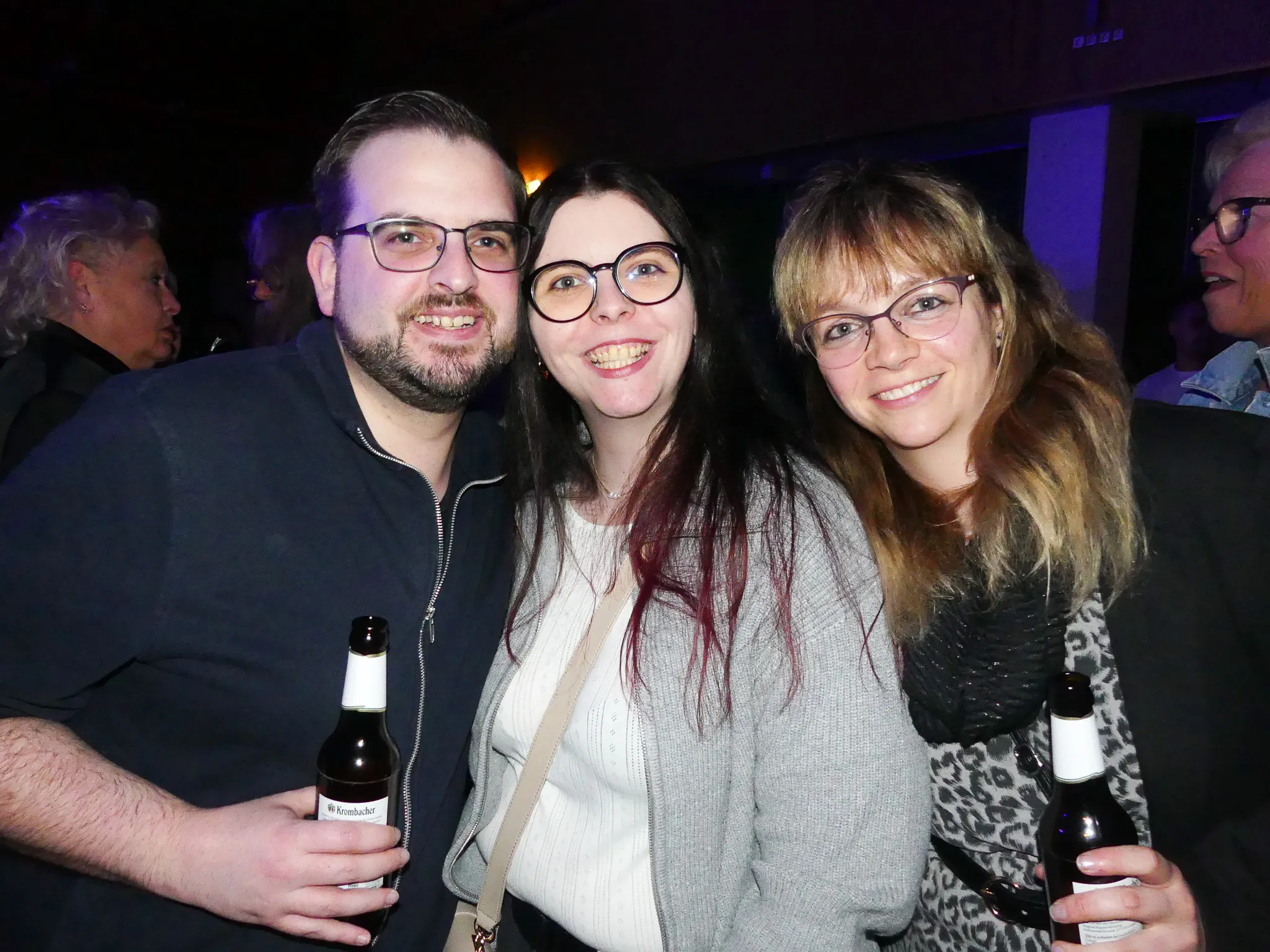 Drei lächelnde Menschen posieren eng zusammen auf einer Party, jeder hält eine Bierflasche. Im Hintergrund sind weitere Partygäste zu sehen.
