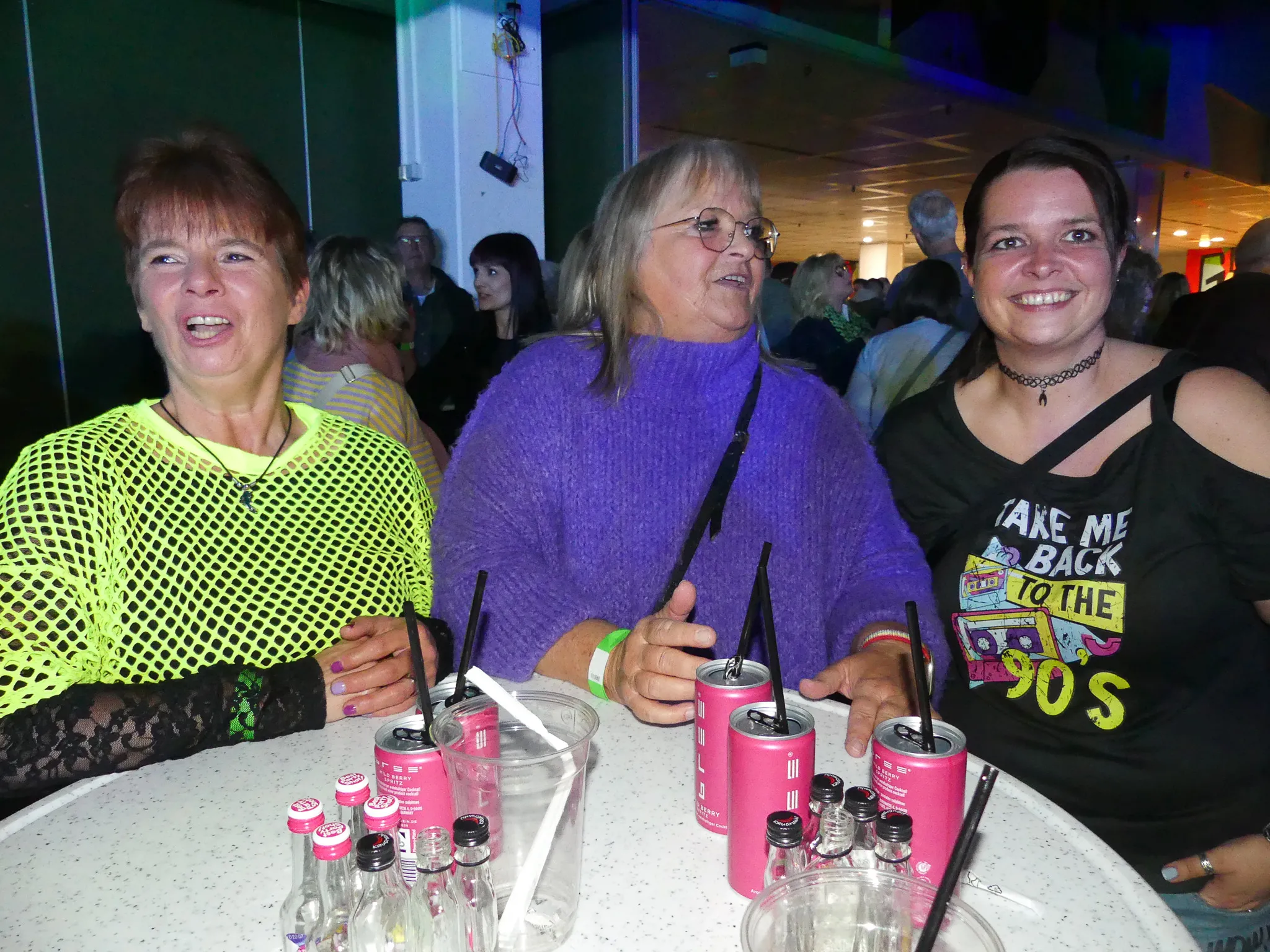 Drei Frauen lächeln in die Kamera, während sie an einem Tisch mit Getränkedosen stehen. Im Hintergrund tanzen weitere Menschen auf einer bunt beleuchteten Tanzfläche.