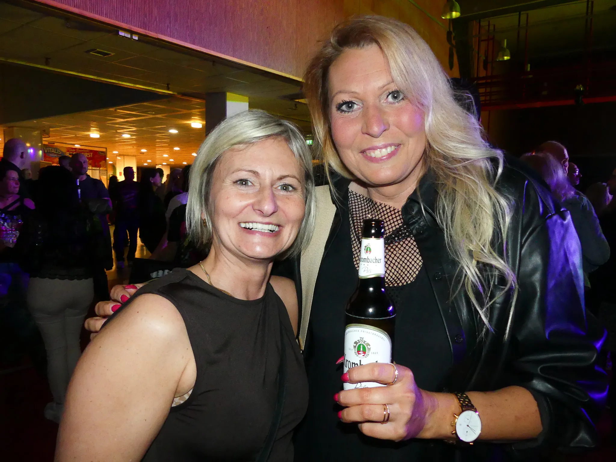 Zwei lächelnde Frauen umarmen sich auf der Tanzfläche einer 80er-/90er-Party, eine von ihnen hält ein Bier in der Hand.