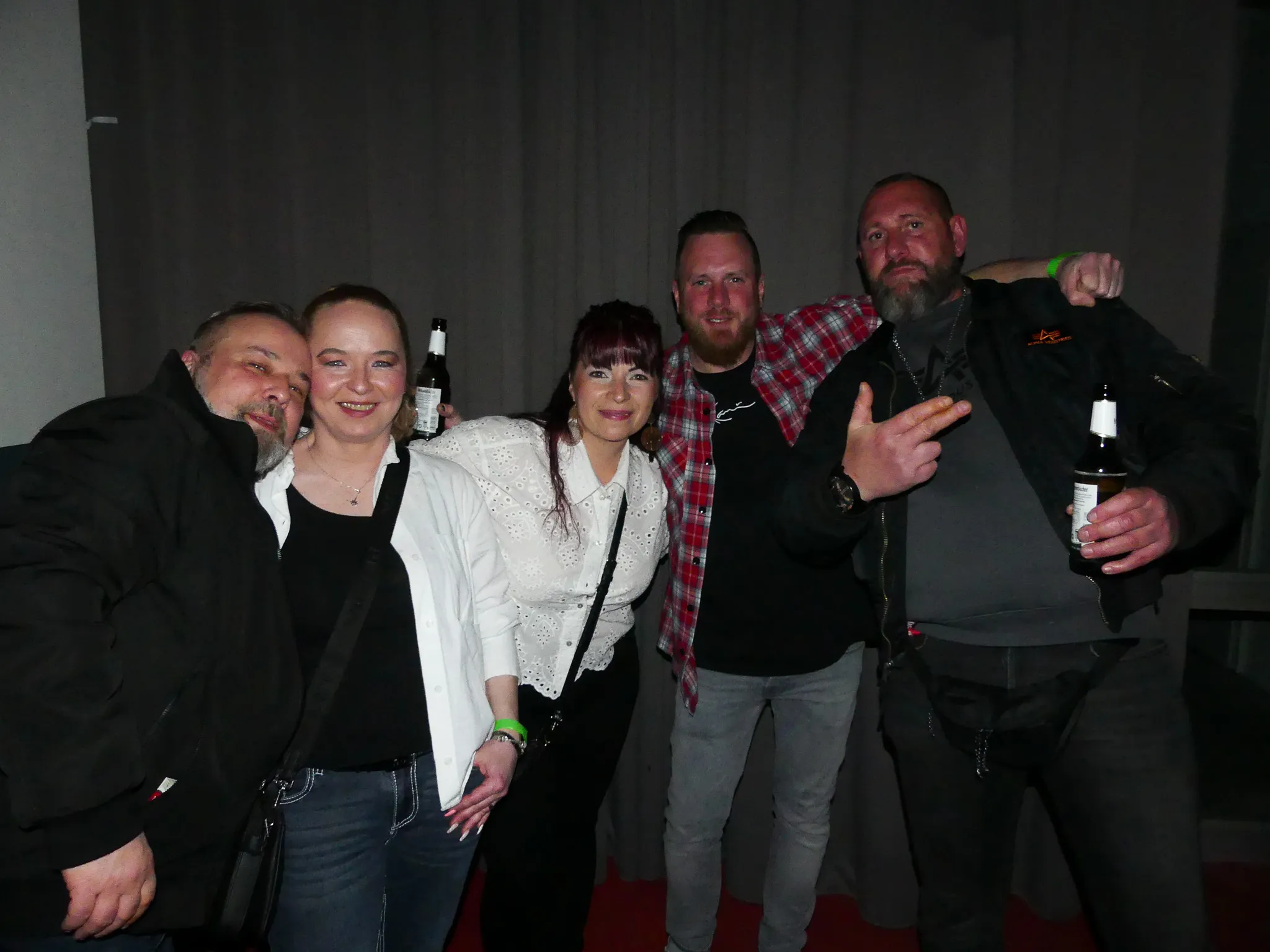 Fünf lächelnde Menschen umarmen sich auf einer Tanzfläche. Sie halten Getränke in der Hand und tragen Freizeitkleidung.