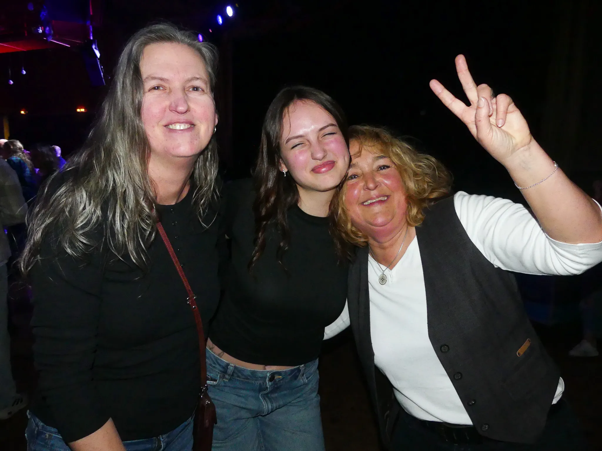 Drei lächelnde Frauen posieren eng zusammen auf einer Tanzfläche, eine von ihnen zeigt das Victory-Zeichen.