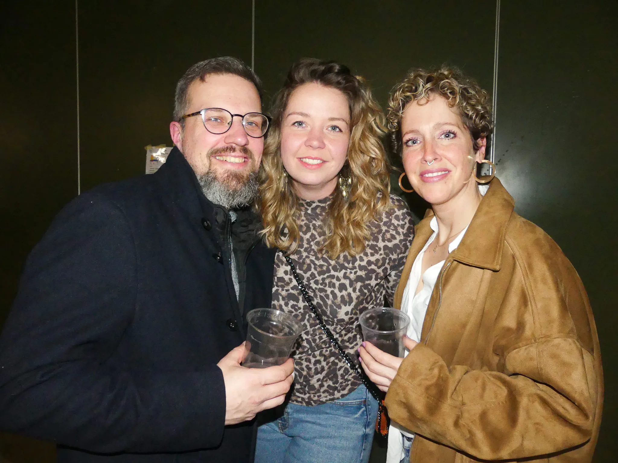 Drei lächelnde Menschen stehen eng zusammen, jeder hält ein Getränk in der Hand. Im Hintergrund eine dunkle Wand.