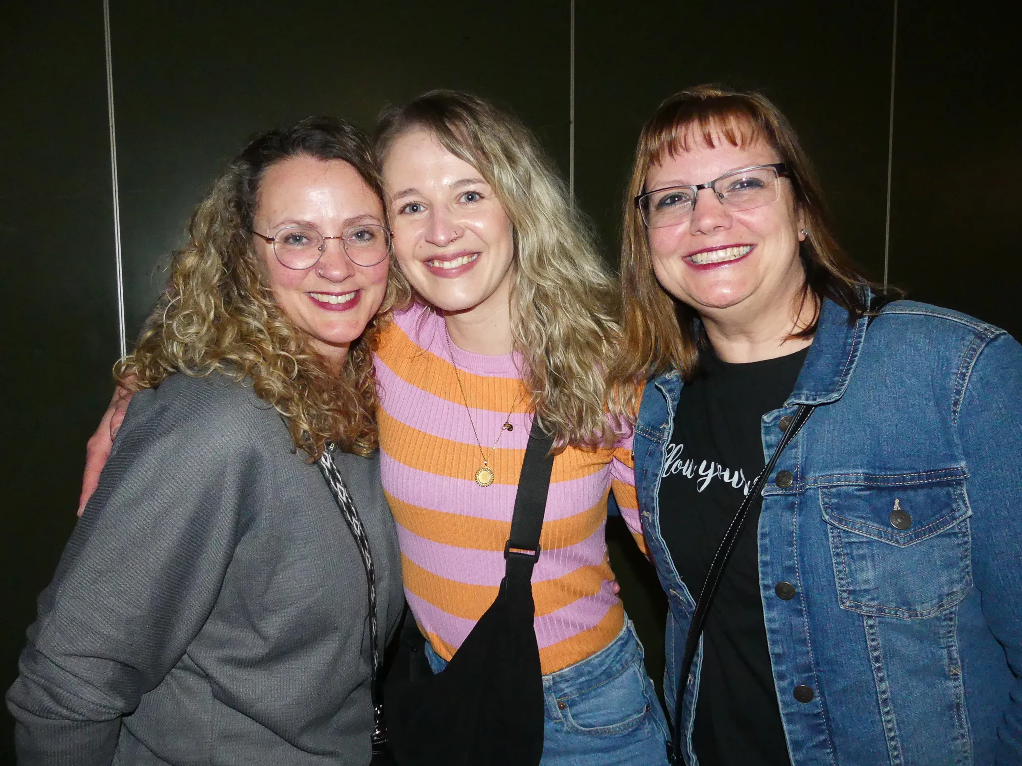 Drei lächelnde Frauen umarmen sich vor einem dunklen Hintergrund. Die linke Frau trägt ein graues Jackett, die mittlere einen pink-lila gestreiften Pullover und die rechte eine Jeansjacke.