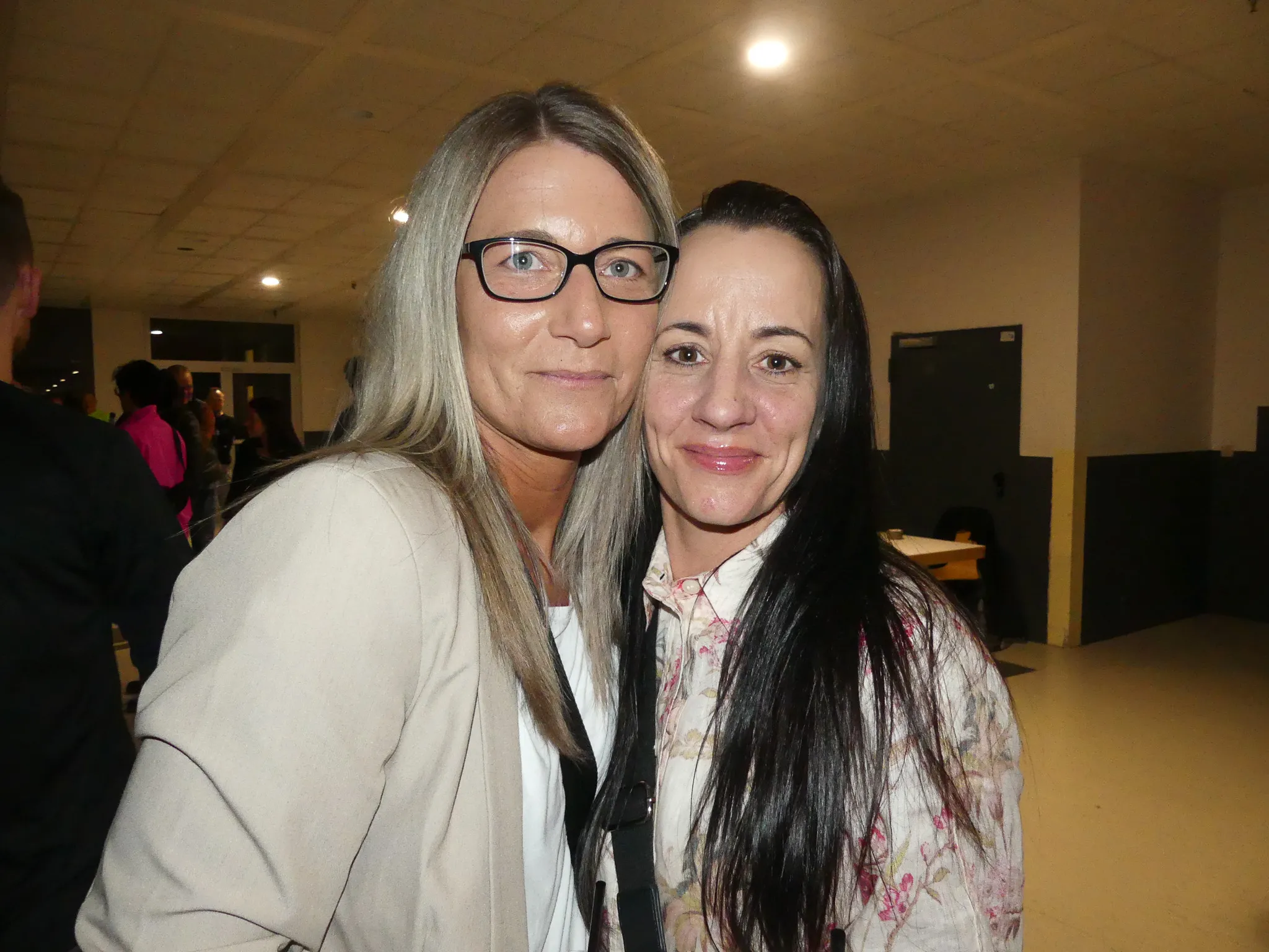 Zwei Frauen umarmen sich vor einer Gruppe von Menschen auf einer Party. Die Frau links trägt eine beige Jacke und die rechte eine Bluse mit Blumenmuster.