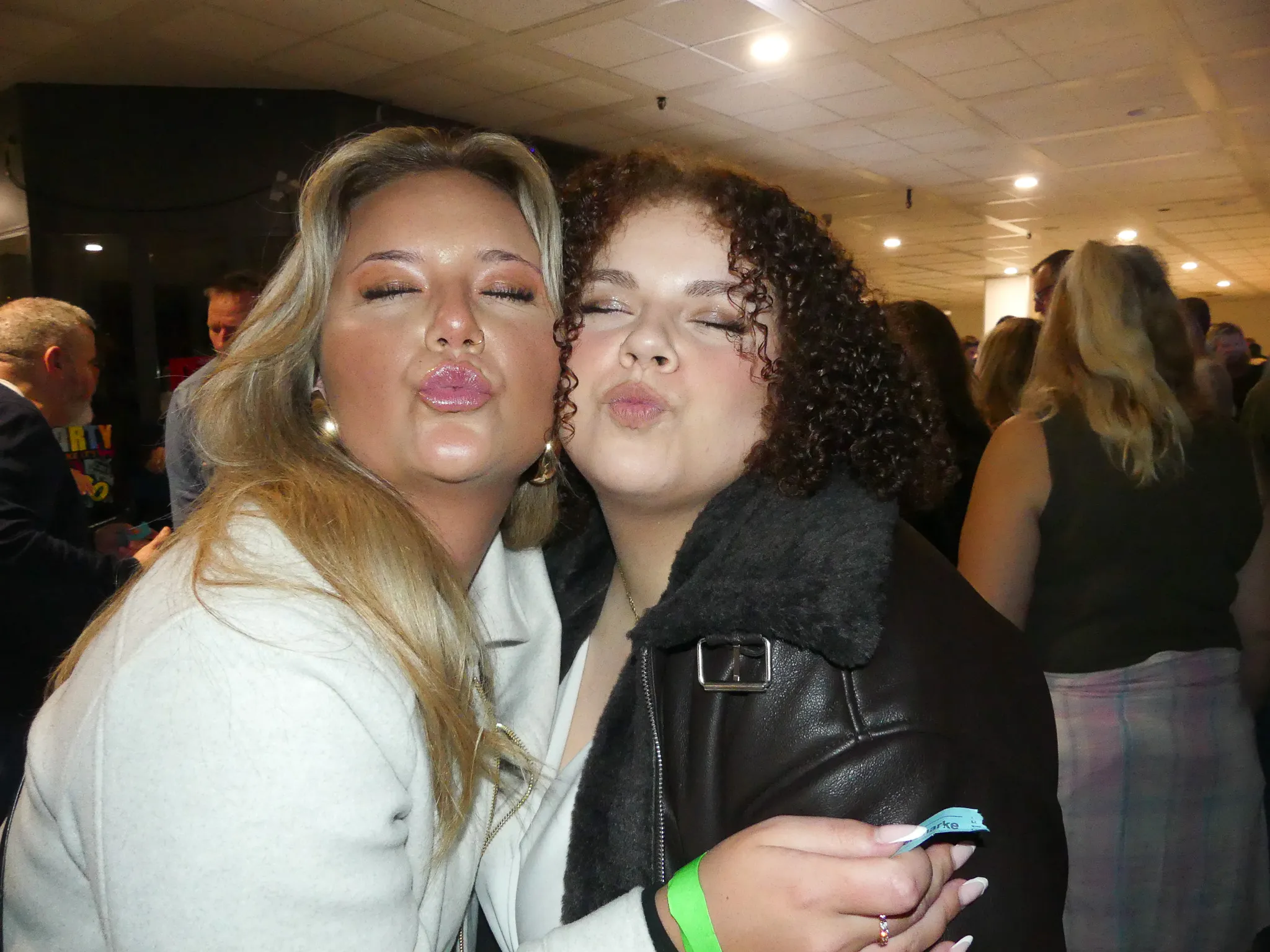 Zwei junge Frauen mit geschlossenen Augen machen Kussgeräusche und umarmen sich auf einer Party. Im Hintergrund sind weitere tanzende Menschen zu sehen.