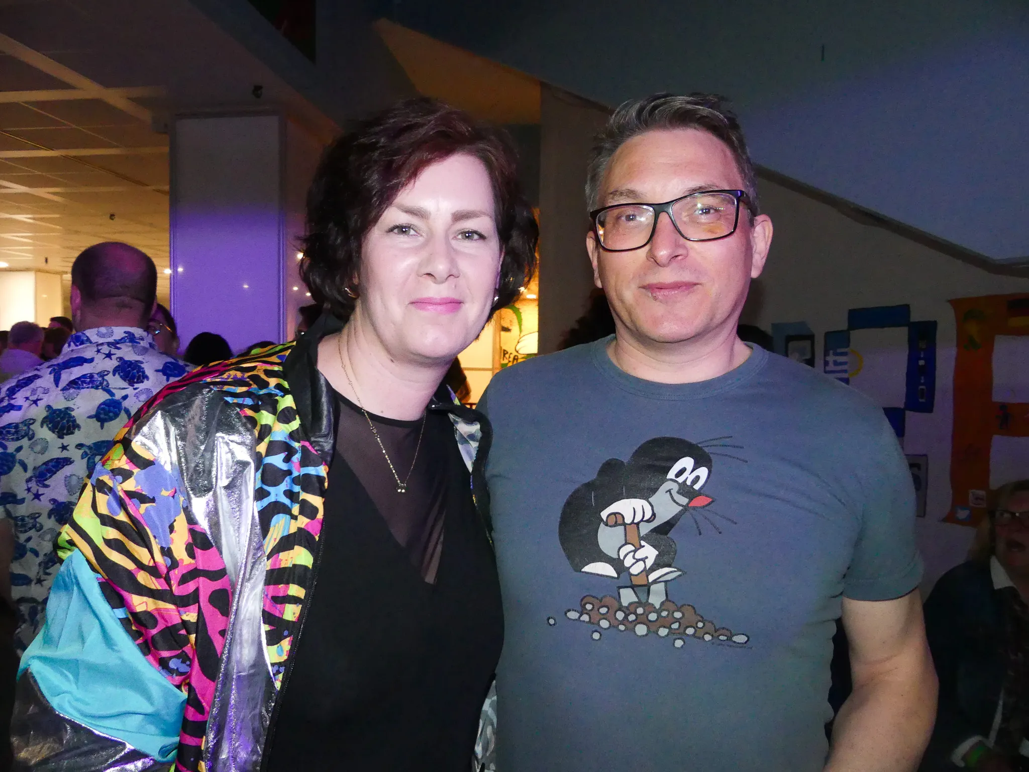 Zwei lächelnde Menschen, eine Frau mit lockigem Haar und einer bunten Jacke sowie ein Mann mit Brille und einem T-Shirt mit Cartoon-Motiv, posieren eng nebeneinander auf einer Party.
