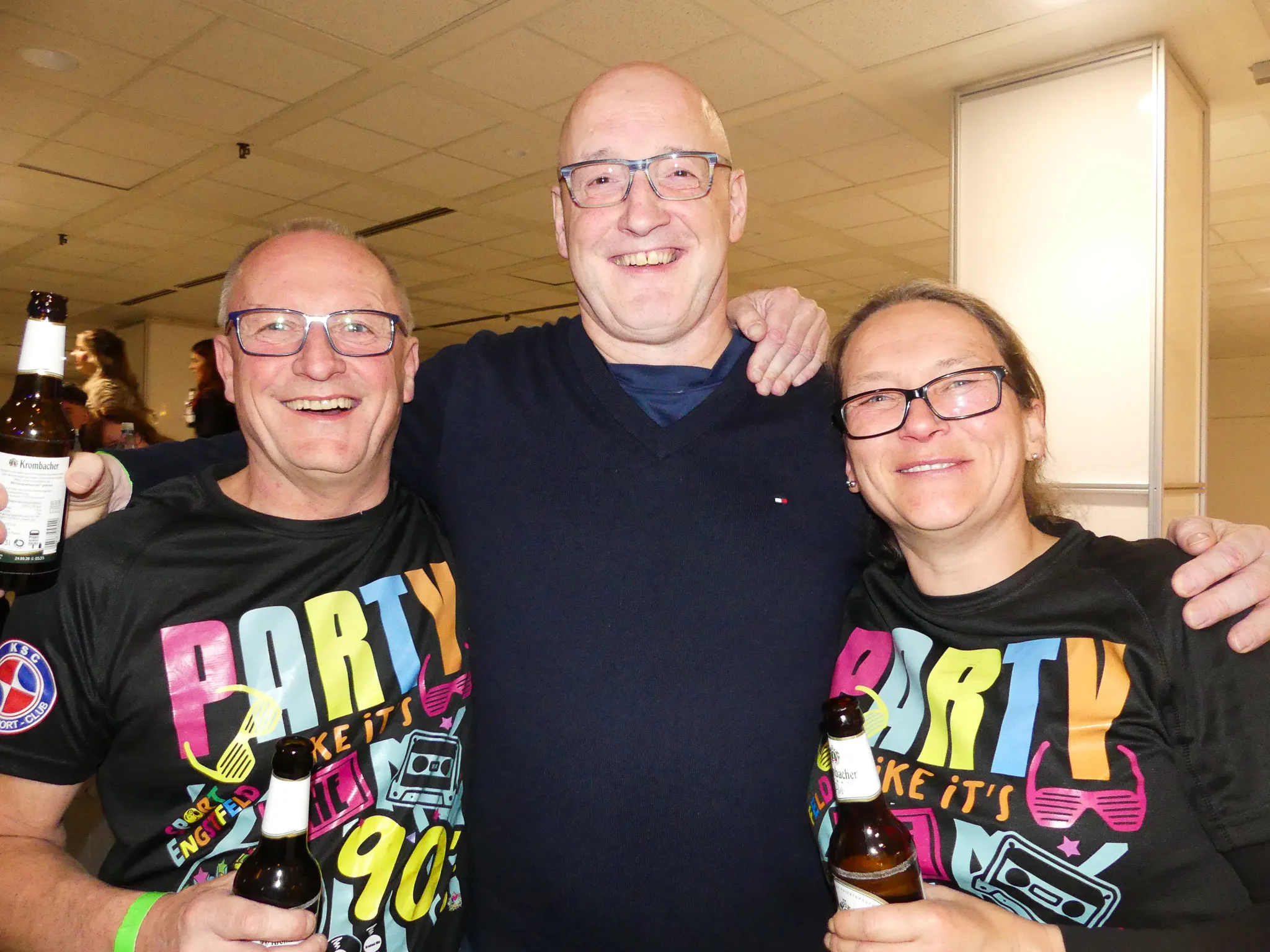 Drei lächelnde Menschen im Alter von etwa 40 bis 60 Jahren stehen eng zusammen. Sie tragen T-Shirts mit dem Aufdruck „Party like it’s…“ und halten Bierflaschen in der Hand.
