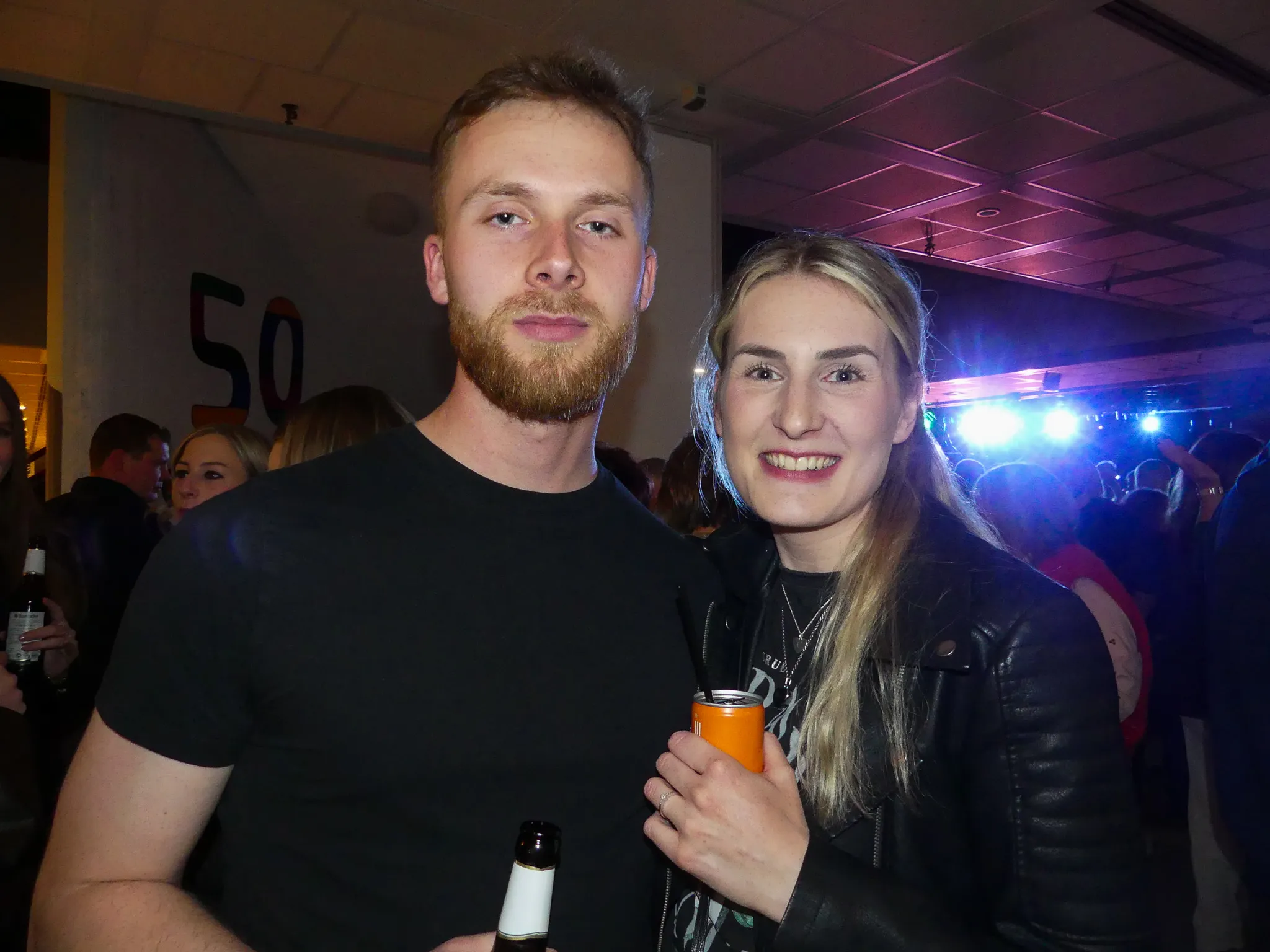 Ein Mann mit Bart und eine Frau mit blonden Haaren stehen lächelnd vor der Kamera, im Hintergrund eine tanzende Menschenmenge und eine große Zahl 50.