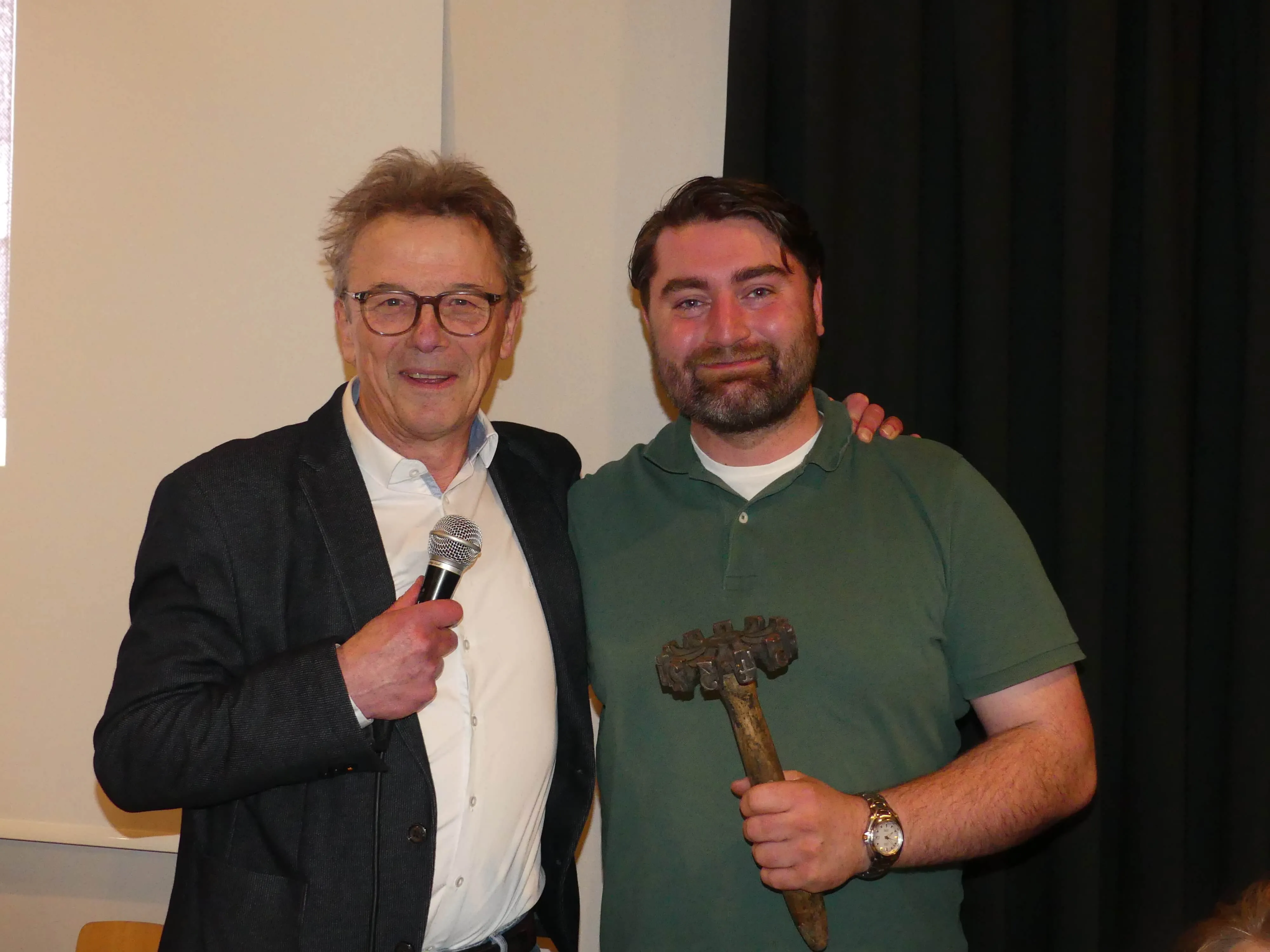 Zwei Männer posieren gemeinsam, der ältere hält ein Mikrofon, der jüngere einen kunstvoll geschnitzten Wanderstab.