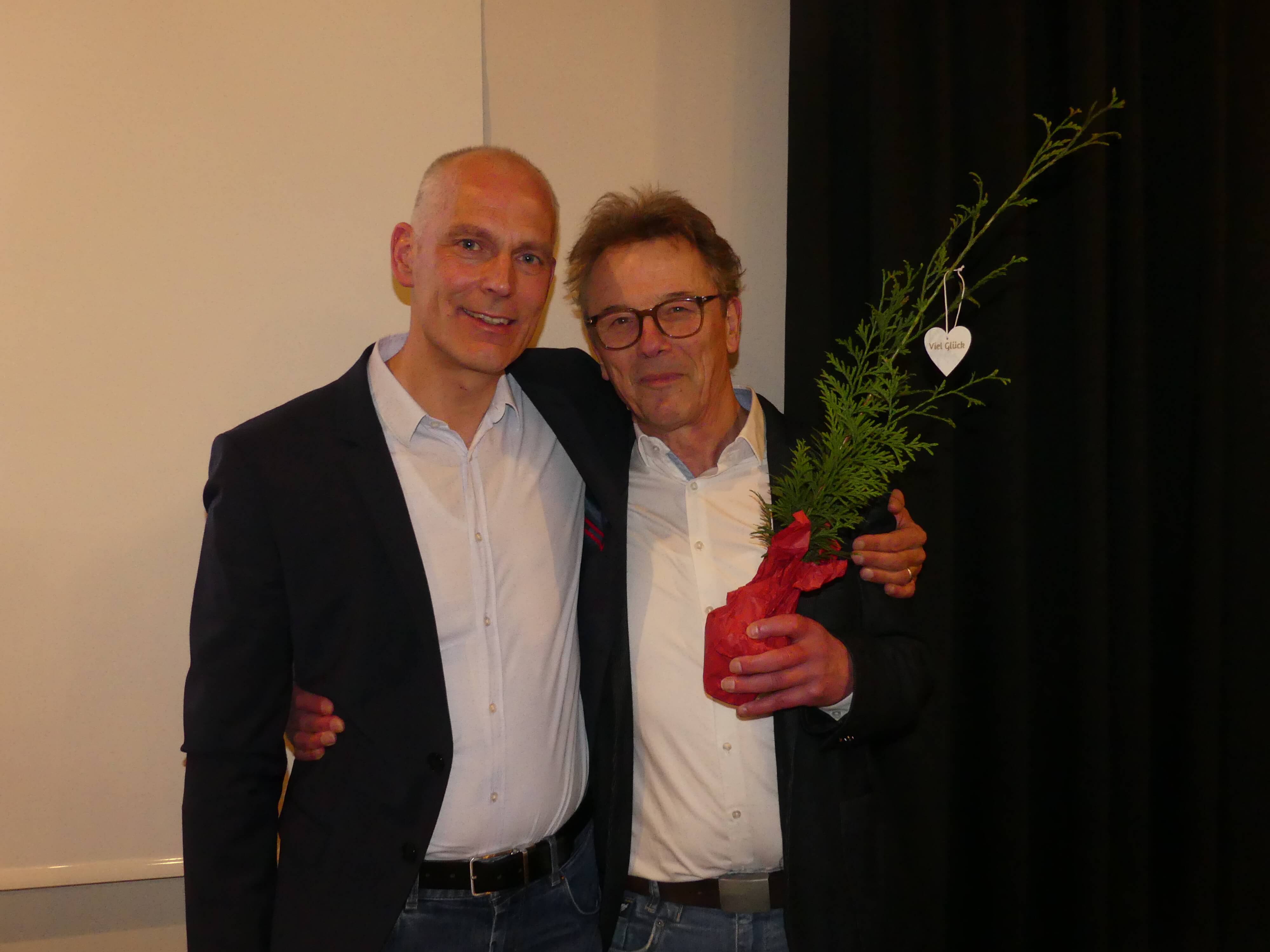 Zwei Männer umarmen sich. Der linke Mann ist glatzköpfig und trägt einen Anzug, der rechte Mann trägt eine Brille und ein Hemd. Er hält einen kleinen Baum im Topf mit einer weißen Herzanhänger.