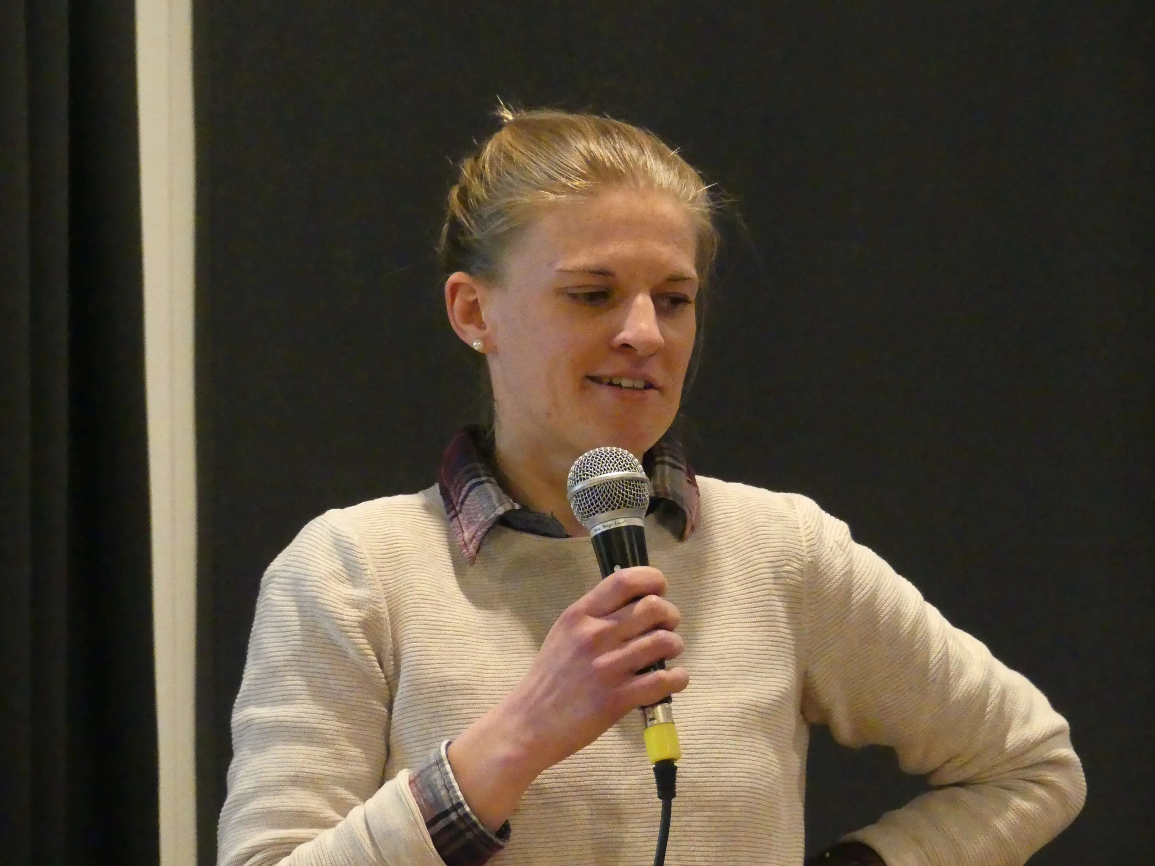Miriam Franzke hält ein Mikrofon und spricht auf einer Veranstaltung. Sie trägt einen grauen Pullover über einem karierten Hemd.