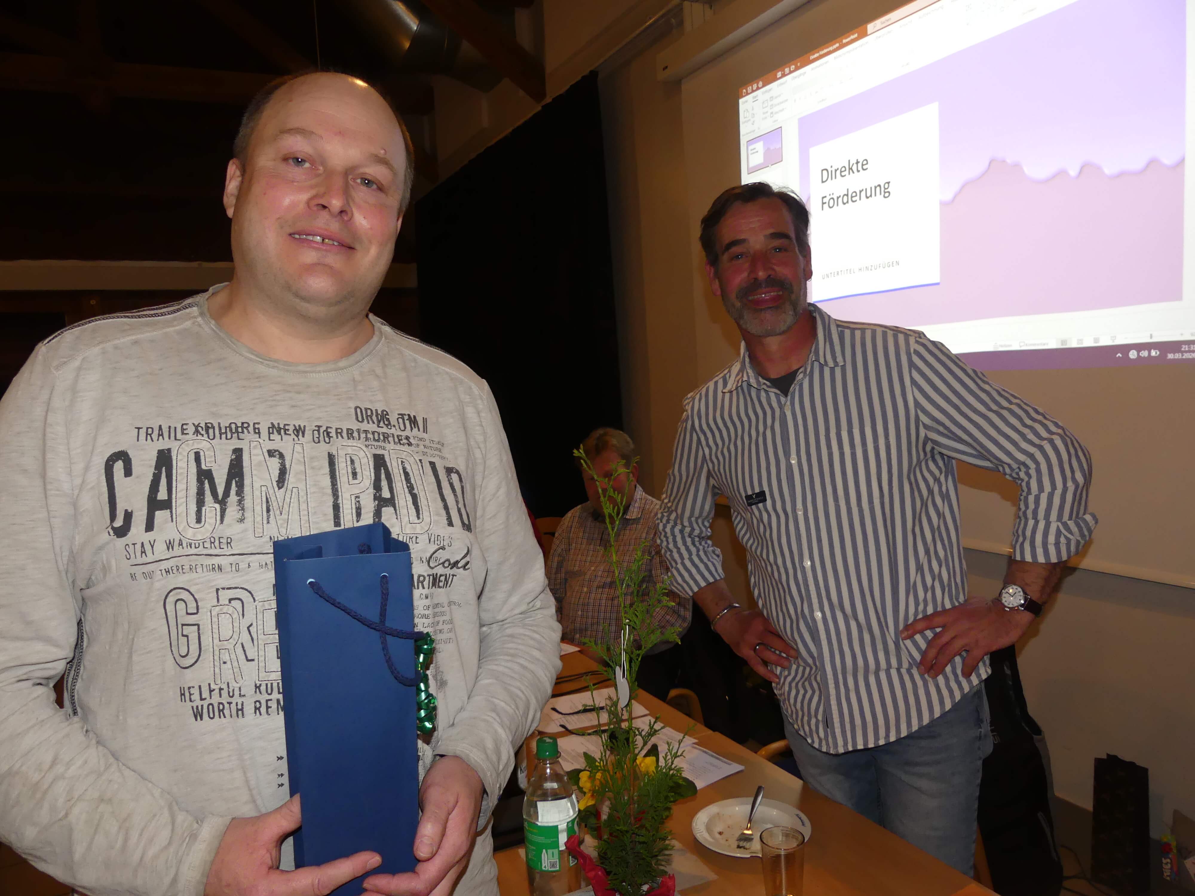 Zwei Männer und eine Frau stehen vor einer Präsentation mit dem Titel „Direkte Förderung“; der Mann links hält einen blauen Ordner.