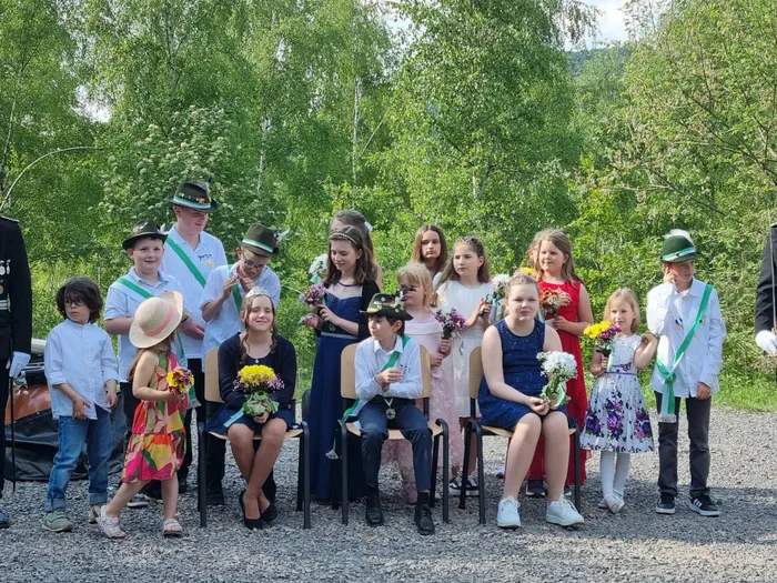 Eine Gruppe von Kindern und Jugendlichen in festlicher Kleidung, einige mit grünen Bändern und traditionellen Hüten, posiert für ein Foto. Im Vordergrund hält ein Mädchen einen Blumenstrauß.