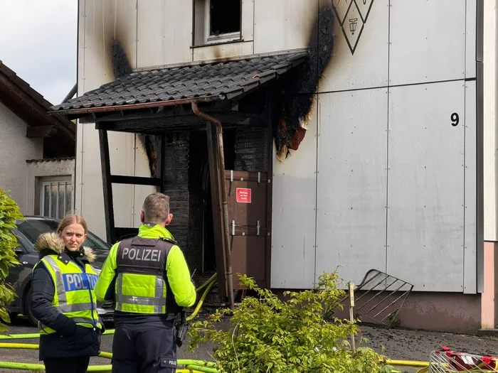 Beschädigte Haustür und Fassade eines Wohnhauses nach einem Brand, vor dem zwei Polizisten stehen. Im Hintergrund ein rotes Verbotsschild und ein umgestürztes Metallgitter.