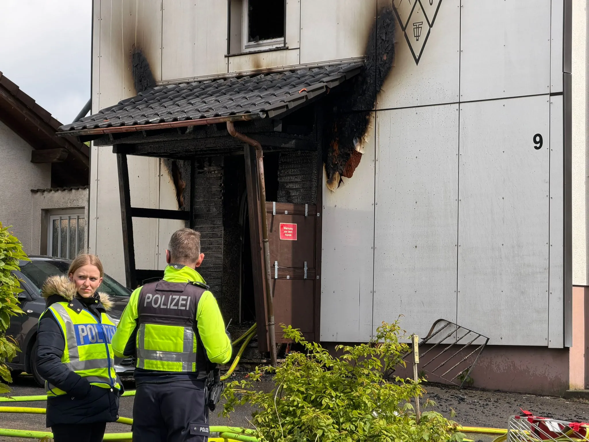 Beschädigte Haustür und Fassade eines Wohnhauses nach einem Brand, vor dem zwei Polizisten stehen. Im Hintergrund ein rotes Verbotsschild und ein umgestürztes Metallgitter.