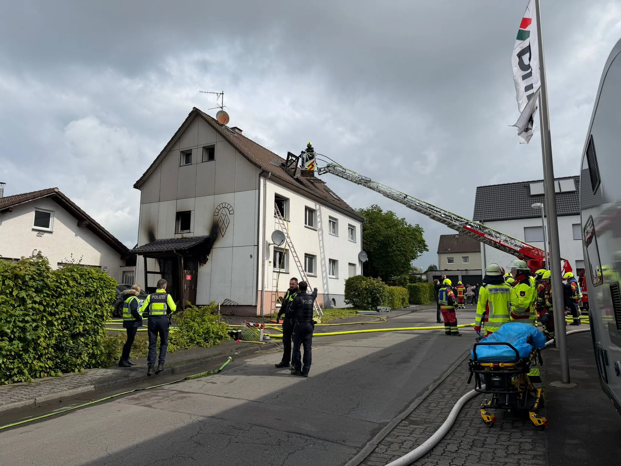 Feuerwehrleute bekämpfen einen Brand in einem zweigeschossigen Wohnhaus, während Polizisten die Einsatzstelle absichern.