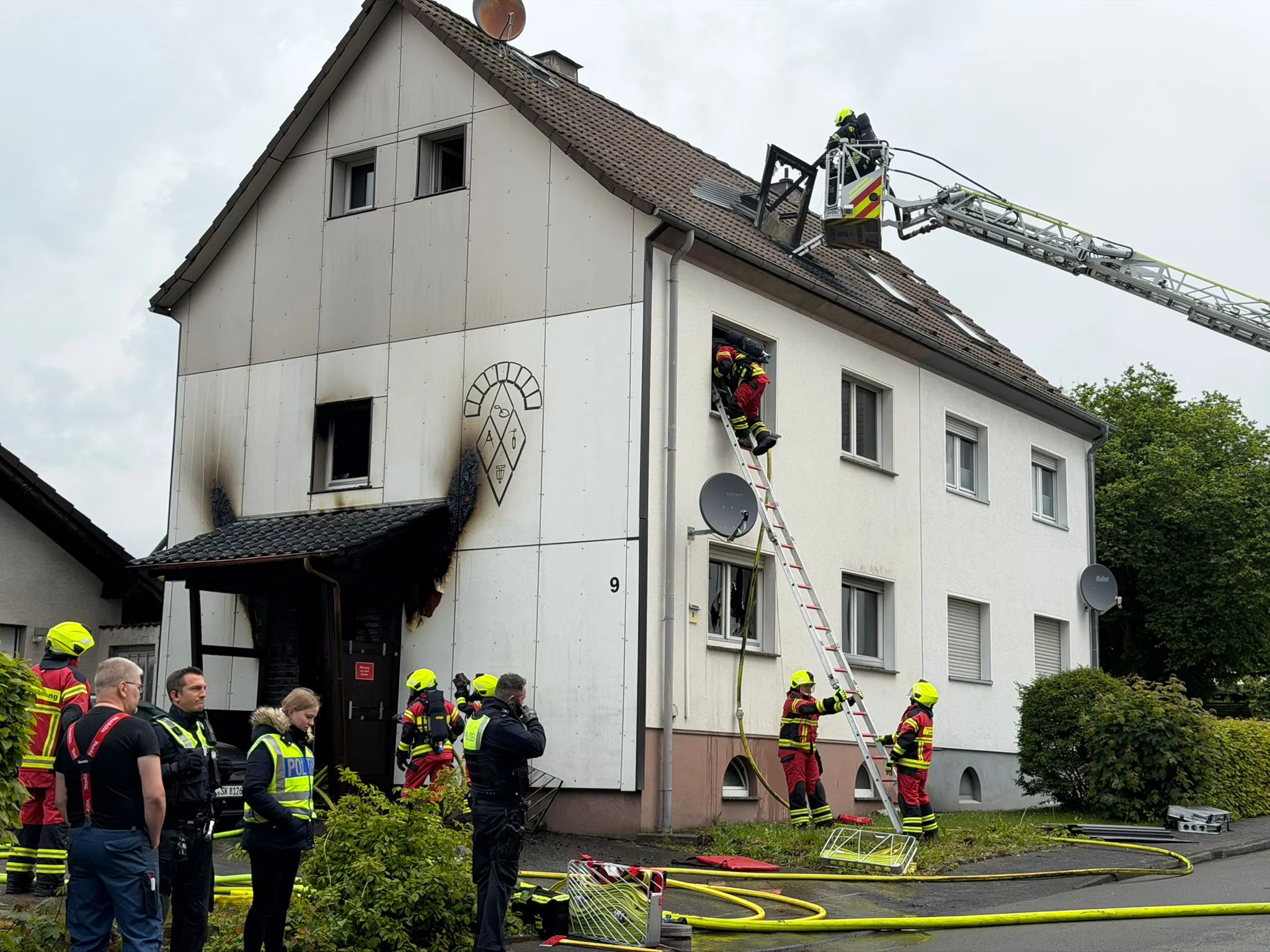 Feuerwehrleute bekämpfen einen Brand in einem zweigeschossigen Wohnhaus mit einer Drehleiter und einer Leiter. Einsatzkräfte stehen am Boden und beobachten die Löscharbeiten.