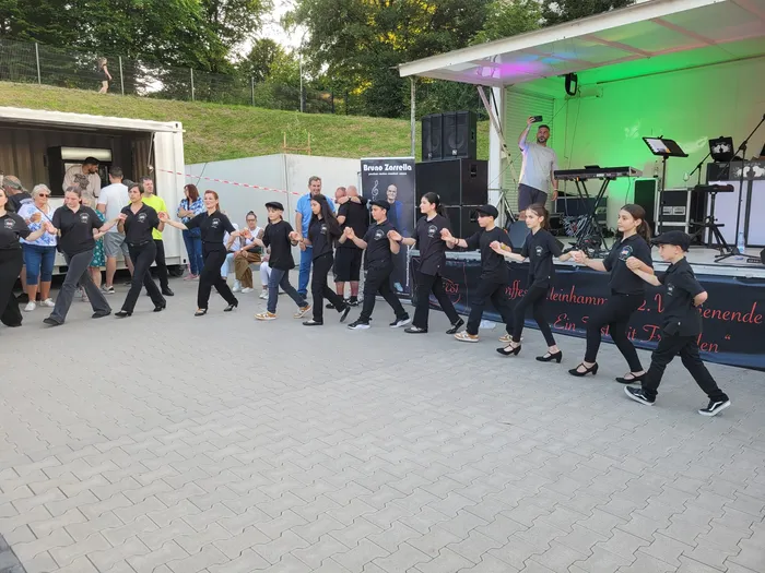 Eine Gruppe von Kindern und Jugendlichen in dunklen Uniformen tanzt auf einem gepflasterten Platz vor einer Bühne. Im Hintergrund steht ein Lastwagen mit der Aufschrift „Bruno Zarrella“. Menschen stehen und sitzen im Hintergrund und beobachten die Aufführung.
