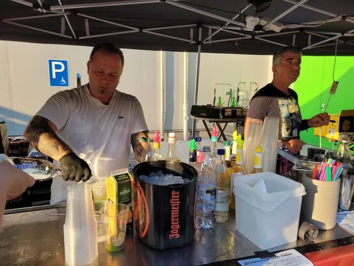 Zwei Männer bedienen eine Bar unter einem weißen Zelt. Auf der Theke stehen diverse Spirituosen und Gläser, im Hintergrund ein Parkplatzschild.