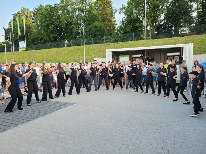 Eine große Gruppe von Menschen tanzt in einer Reihe auf einem gepflasterten Platz. Die Tänzer tragen größtenteils dunkle Kleidung. Im Hintergrund steht ein Wohnwagen und eine grüne Hügelkuppe.