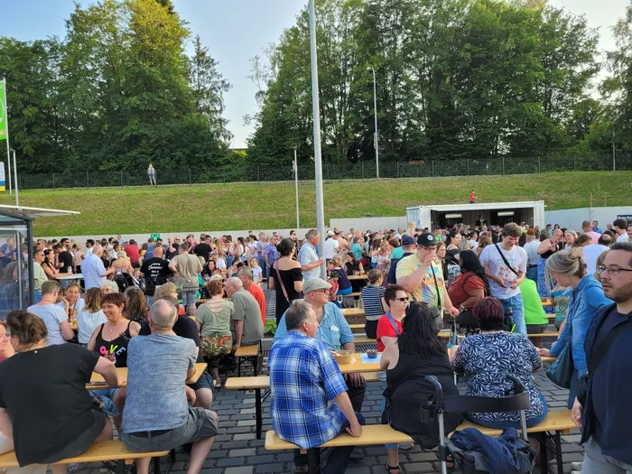 Eine große Menschenmenge sitzt und steht an Biertischgarnituren vor einem Veranstaltungsort im Freien. Im Hintergrund ist eine grüne Hügellandschaft zu sehen.