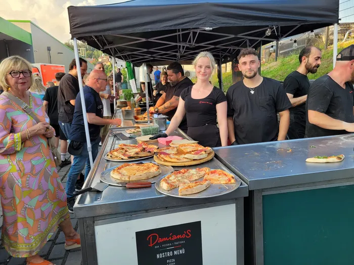 People are gathered around a food stand with several pizzas on display. A woman in a patterned shirt stands to the left, while staff members prepare and serve food from behind the counter. The stand is labeled 