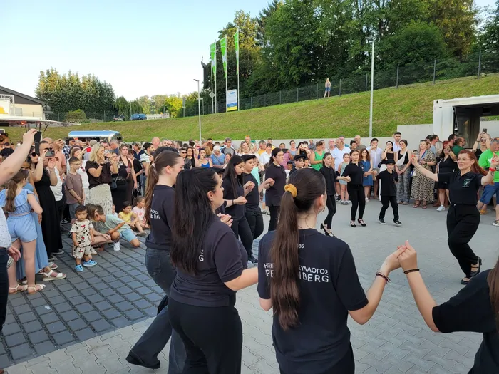 Eine Gruppe von Menschen steht und sitzt auf einem gepflasterten Platz vor einem Gebäude. Im Vordergrund tanzen drei Frauen in dunklen T-Shirts im Kreis, umgeben von Zuschauern. Im Hintergrund sind Bäume und grüne Hügel zu sehen.