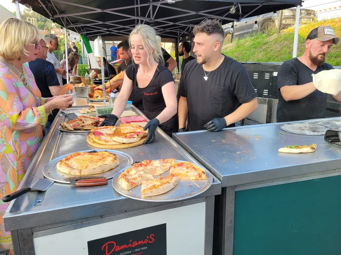 Mitarbeiter von Damiano's Pizzeria bereiten vor Kunden an einem Stand Pizza zu. Mehrere Pizzen liegen auf dem Tisch, eine wird belegt.