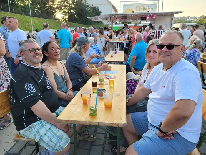 Menschen sitzen an langen Biertischbänken und genießen Getränke und Gespräche bei einem Sommerfest im Freien. Im Hintergrund ist ein Gebäude mit der Aufschrift „Bitourger“ zu sehen.