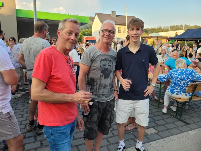 Drei Männer posieren im Freien, umgeben von einer Menschenmenge bei einer Veranstaltung. Der Mann links trägt ein rotes T-Shirt, der in der Mitte ein graues T-Shirt mit einem Porträt und der rechte Mann ein blaues Poloshirt. Sie halten alle Gläser in der Hand. Im Hintergrund sind weitere Menschen und Zelte zu sehen.