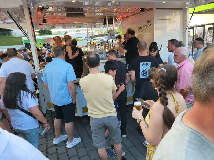 Menschen stehen an einem Getränkestand mit der Aufschrift „Bitte ein Bier“ an einem belebten Platz. Viele Besucher genießen den Sommerabend und die mediterrane Atmosphäre.
