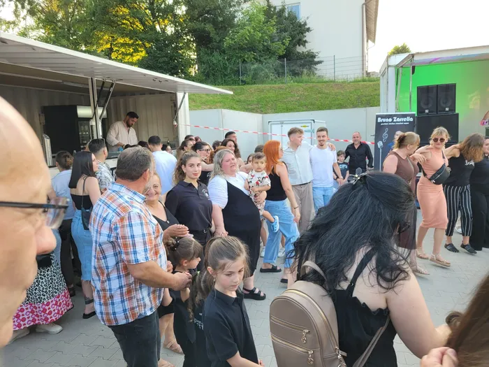 Eine große Menschenmenge steht vor einem Foodtruck und genießt die Veranstaltung „Griechenland trifft Italien“. Im Hintergrund sind weitere Besucher und Lautsprecheranlagen zu sehen.