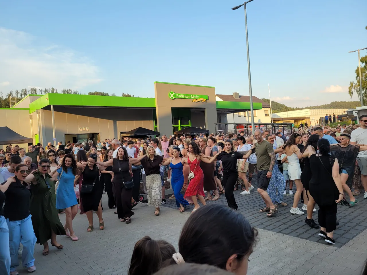 Eine große Menschenmenge tanzt im Freien vor einem Supermarkt. Im Hintergrund sind Bäume und ein bewölkter Himmel zu sehen.