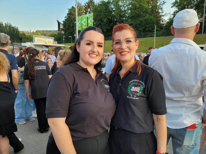 Zwei lächelnde Frauen mit dunklen und roten Haaren stehen eng nebeneinander vor einer Menschenmenge. Beide tragen dunkelblaue Poloshirts mit dem Aufdruck „Plettenberg“ und einem Logo mit griechischen Motiven. Im Hintergrund sind Bäume und Gebäude zu sehen.