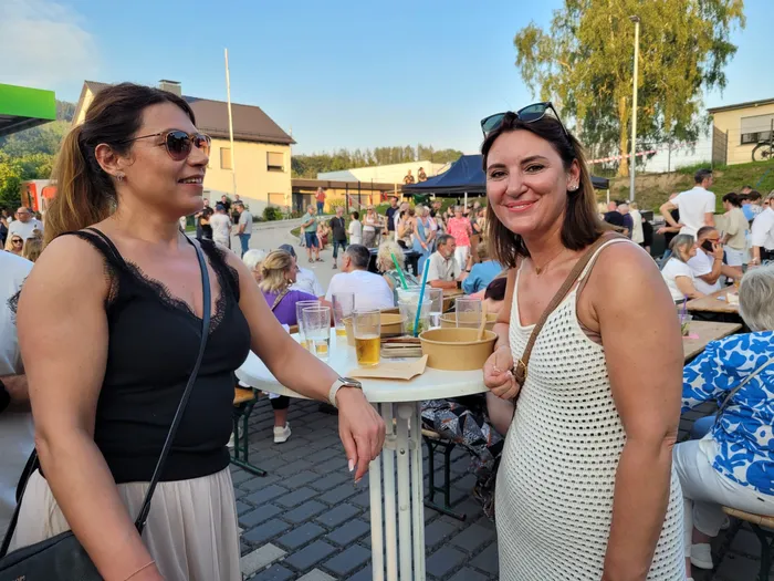 Zwei lächelnde Frauen stehen vor einem Tisch mit Getränken im Freien, im Hintergrund eine Menschenmenge und ein Gebäude mit blauem Dach. Das Bild zeigt eine lebhafte Veranstaltung mit vielen Besuchern.