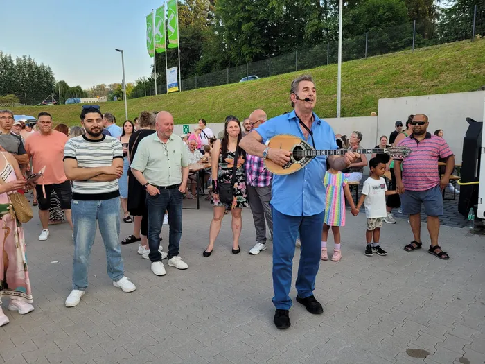 Ein Mann spielt auf einer Laute vor einer Gruppe von Menschen im Freien. Die Zuschauer stehen und sitzen auf einer Fläche mit Grünflächen im Hintergrund.