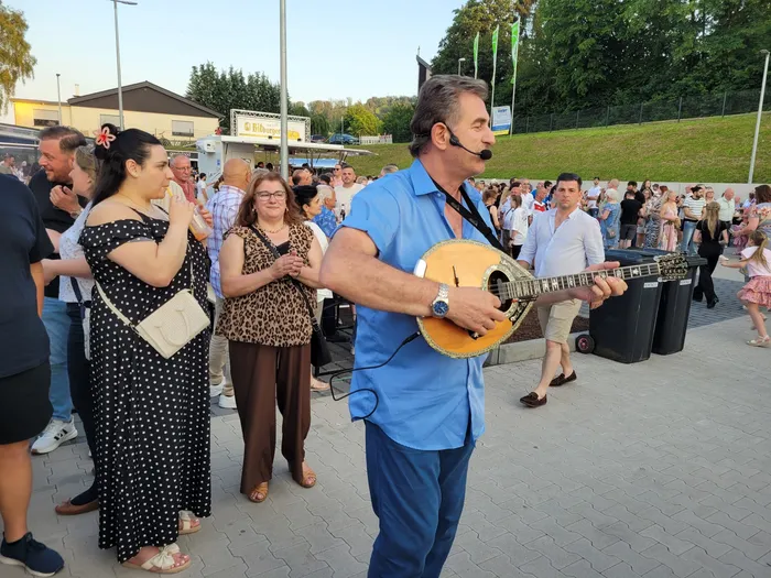 Ein Mann spielt auf einer Mandoline vor einer großen Menschenmenge im Freien. Im Hintergrund sind Gebäude und Bäume zu sehen.