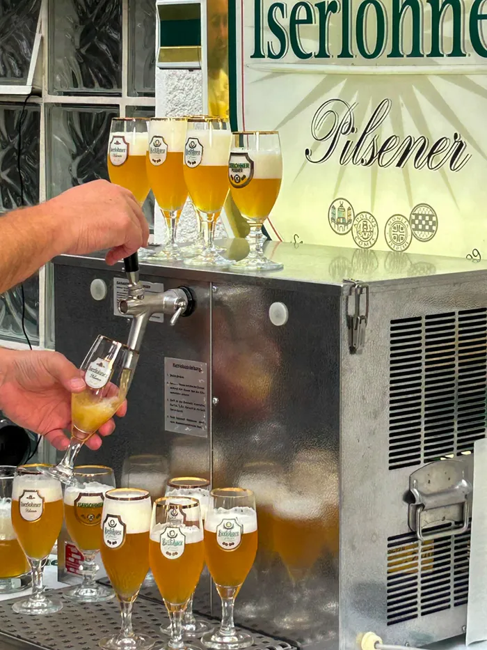 Hände zapfen Iserlohner Pilsener aus einem Zapfhahn in ein Glas, umgeben von weiteren gefüllten Biergläsern.
