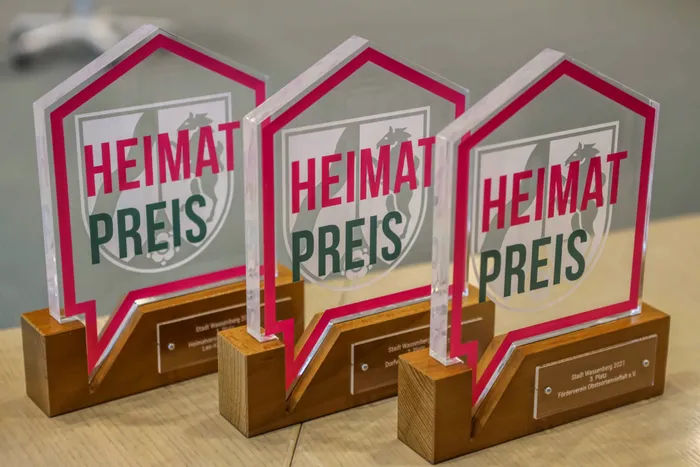 Drei gläserne Pokale mit der Aufschrift „HEIMATPREIS“ stehen auf einem dunklen Untergrund. Die Pokale sind mit einem pinkfarbenen Rahmen versehen und tragen Gravuren mit dem Namen der Stadt Kierspe und dem Jahr 2027.