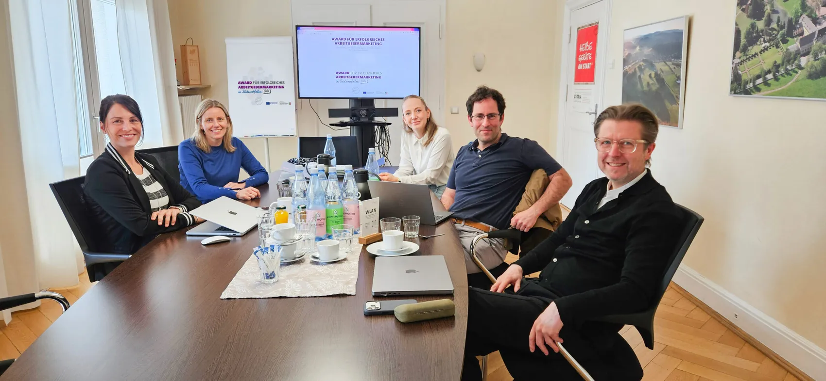 Fünf Personen sitzen an einem Konferenztisch und blicken in die Kamera. Auf dem Tisch stehen Wasserflaschen, Gläser und Teller. Im Hintergrund ist eine Präsentation mit dem Titel „Award für erfolgreiches Arbeitgebermarketing“ zu sehen.