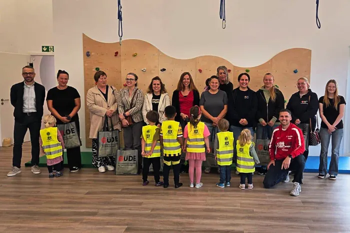 Eine Gruppe von Erwachsenen und Kindern posiert in einem Raum mit einer Kletterwand, wobei einige Kinder gelbe Warnwesten tragen.