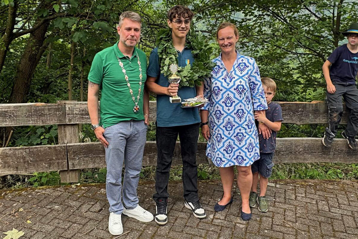 Patrick Hansmann, Nina Schauerte und Ruben Grüber stehen nach dem Kinderprobevogelschießen mit dem Pokal vor einer Holzbarriere. Ein Junge steht im Hintergrund.