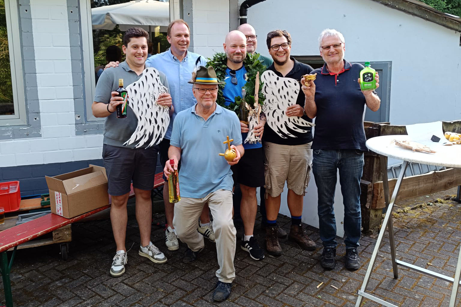 Eine Gruppe von sechs Männern posiert mit erlegten Vögeln und Sachpreisen im Freien. Ein Mann in der Mitte hält eine goldene Insignie.