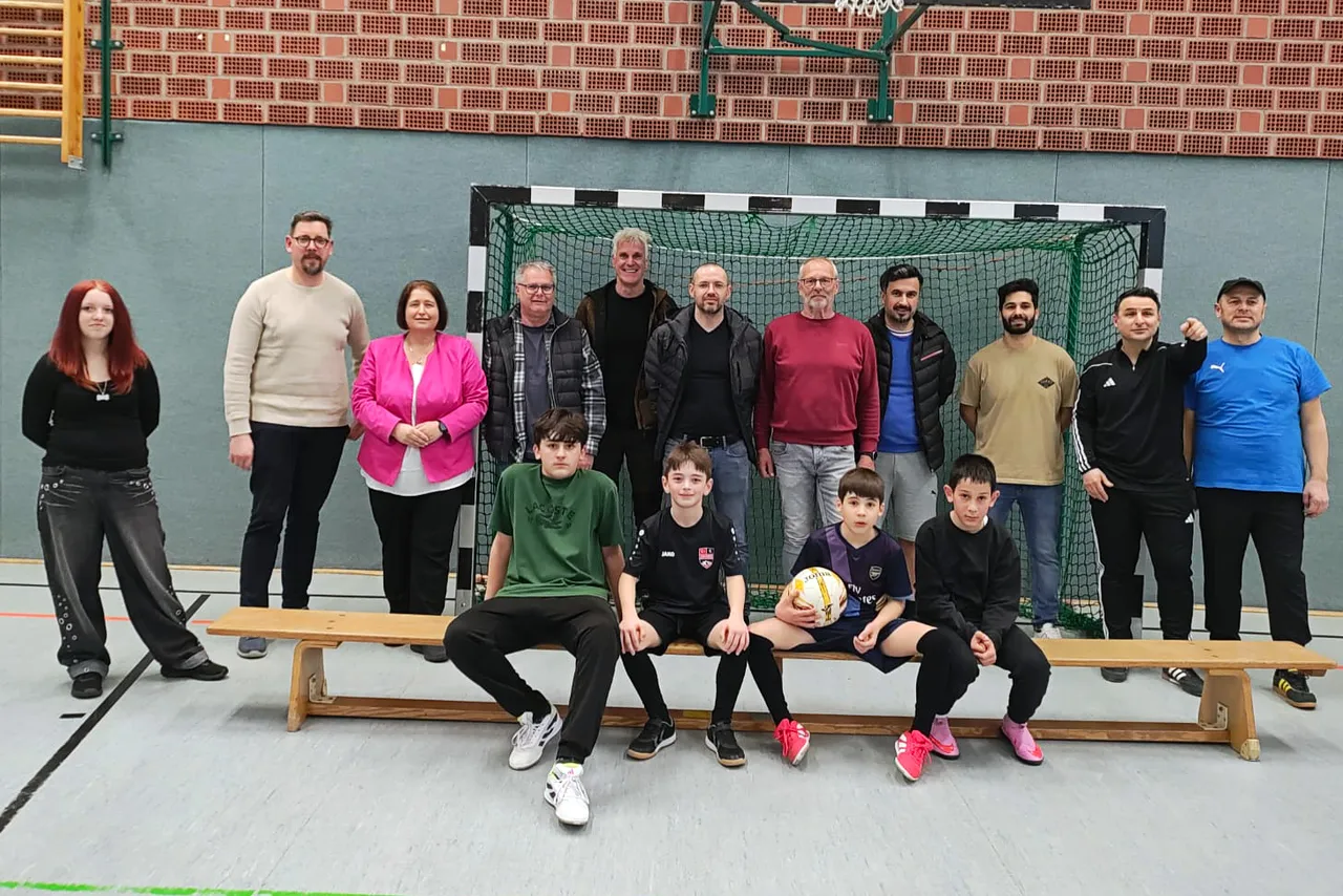 Gruppe von Erwachsenen und Jugendlichen vor einem Fußballtor in einer Sporthalle. Einige halten einen Fußball, andere sitzen oder stehen lächelnd.