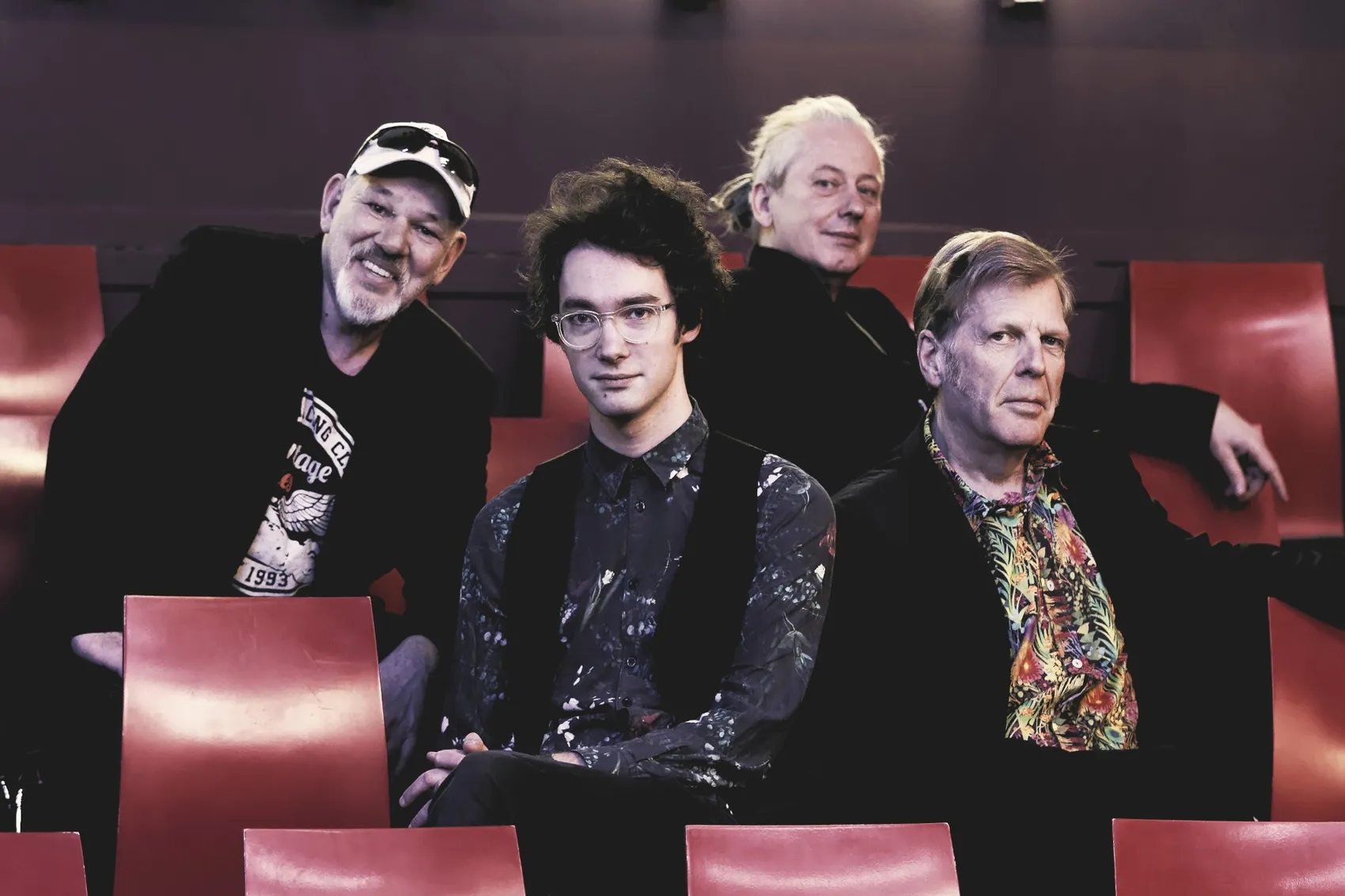 Vier Männer sitzen auf roten Sitzen in einem Theater. Der Mann in der Mitte blickt direkt in die Kamera, die anderen drei sind seitlich abgewandt.