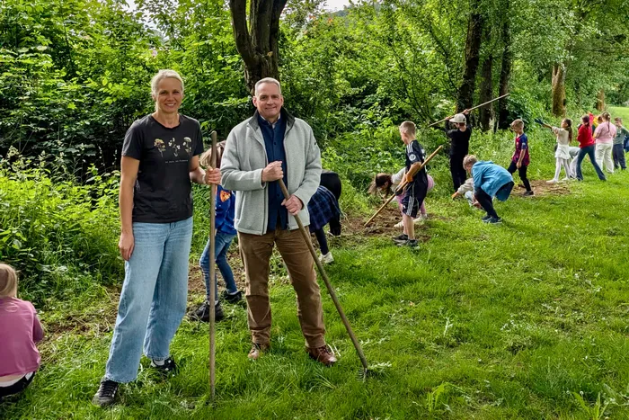 Zwei Erwachsene stehen lächelnd mit Rechen in der Hand auf einer Wiese, im Hintergrund Kinder bei der Arbeit an einem Blühstreifen. Die Frau trägt Jeans und ein T-Shirt, der Mann eine Jacke und Hose.