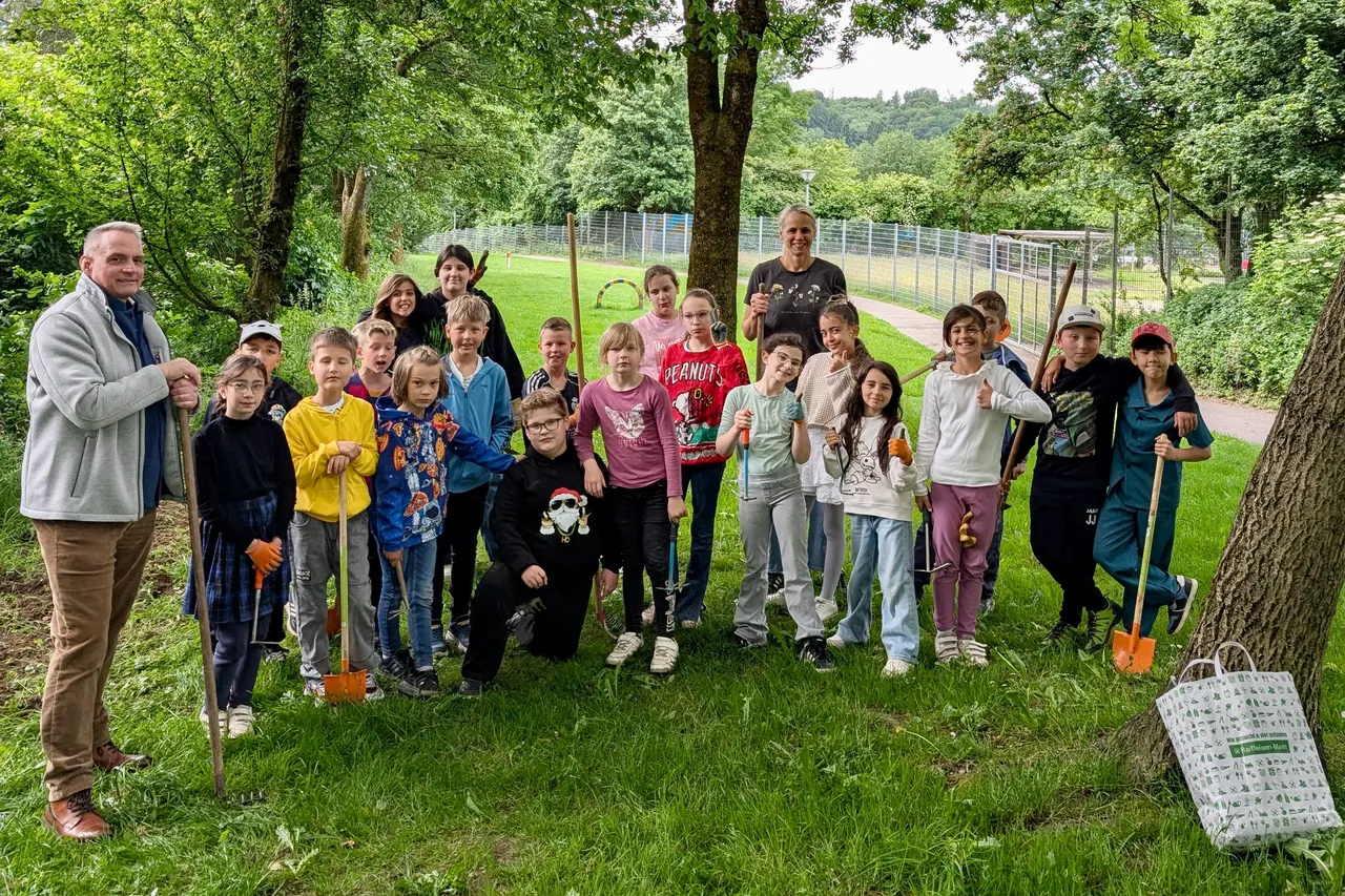 Eine Gruppe von Kindern und zwei Erwachsene steht lächelnd vor einem Blühstreifen. Einige Kinder halten kleine Schaufeln oder Gießkannen in den Händen.