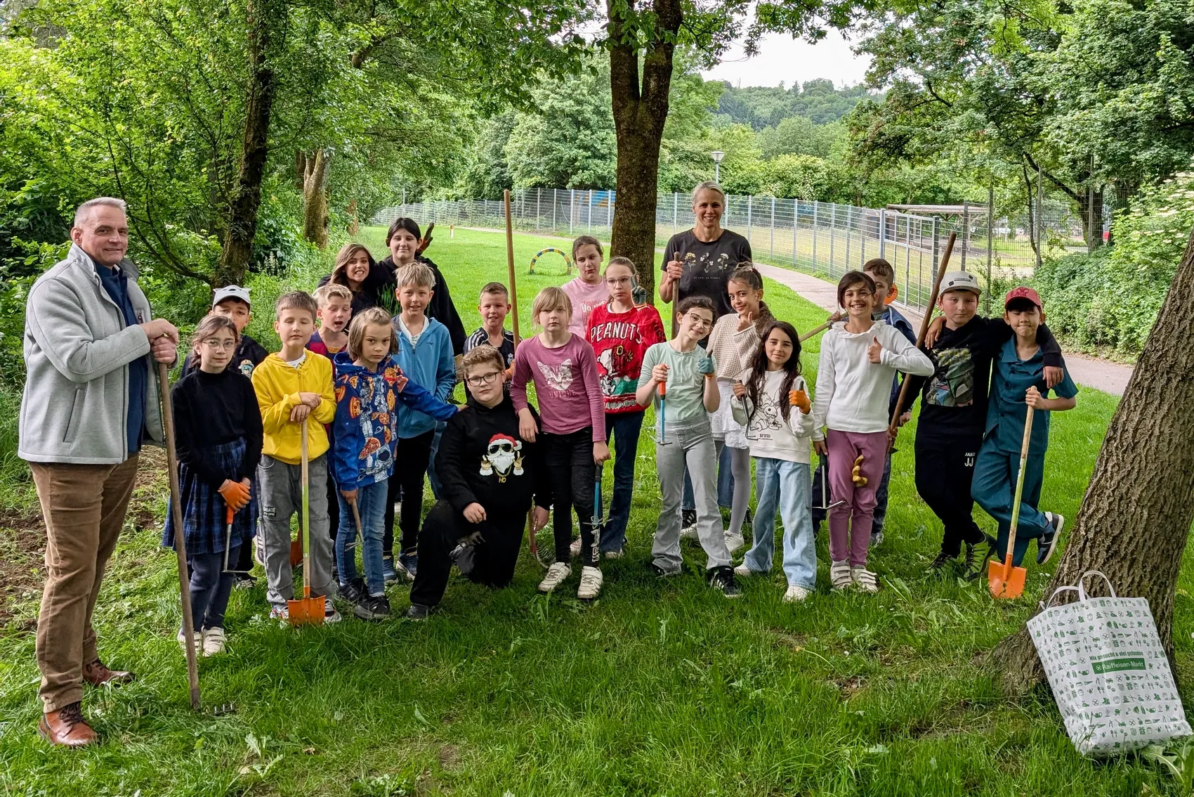 Eine Gruppe von Kindern und zwei Erwachsene steht lächelnd vor einem Blühstreifen. Einige Kinder halten kleine Schaufeln oder Gießkannen in den Händen.